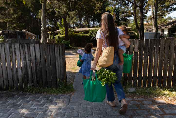 Woman carrying baby and grocery bags with toddler walking in front also carrying a grocery bag
